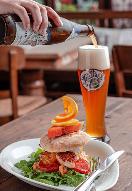 Bier wird in ein Glas gegossen, neben einem gestapelten Hähnchensandwich mit Gemüse auf einem Holztisch.