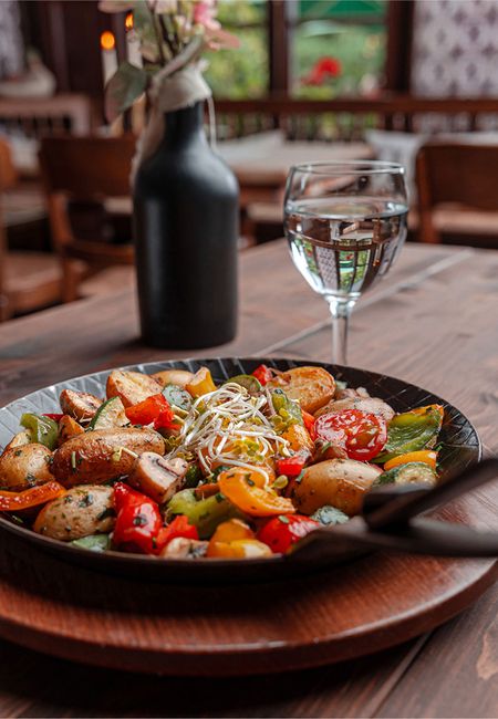 Gebratenes Gemüse in einer Pfanne auf einem Holztisch mit einem Glas Wasser und einer Blumenvase.