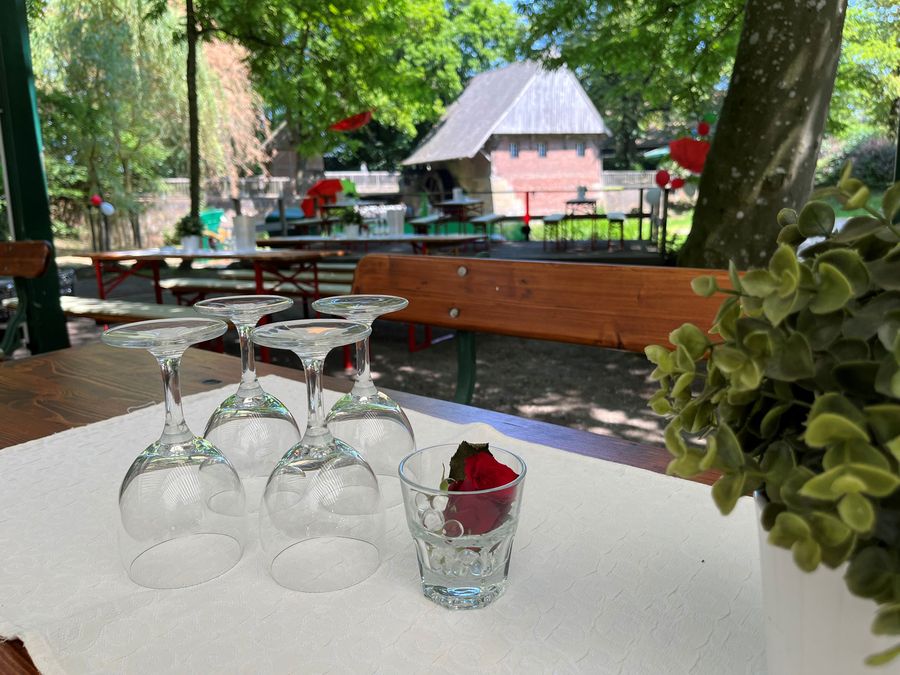 Buitentafel met omgekeerde wijnglazen en een enkele roos, met groene planten en een tuinhuisje op de achtergrond.