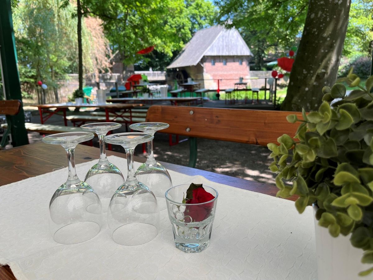 Tisch im Freien mit umgedrehten Weingläsern und einer einzelnen Rose, mit Picknicktischen und Grün im Hintergrund.