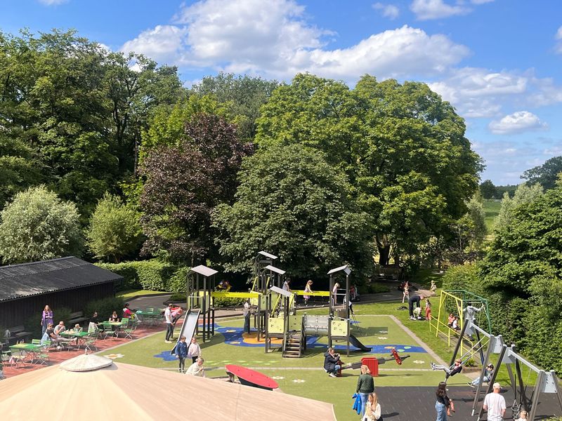 Buitenpark met mensen, speeltoestellen, groene bomen en wat wolken in de lucht.